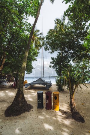 Téléchargez les photos : Un plan vertical grand angle aux Maldives avec poubelles écologiques sur une plage de sable entouré de palmiers, et le bateau en arrière-plan favorise le tourisme durable et la beauté naturelle. - en image libre de droit
