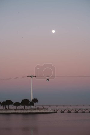 Téléchargez les photos : Vue verticale et discrète d'une belle scène avec une pleine lune, un coucher de soleil rose, la rivière Tejo et un téléphérique. L'effet global est une scène magique sereine et rêveuse capturée à Lisbonne - en image libre de droit