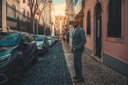Photo for A man in a stylish grey-green suit and sunglasses stands on a narrow street sidewalk, hands in his pockets. Buildings surround him, parked cars line the street, and the sun peeks through at the end. - Royalty Free Image
