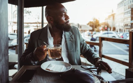 Téléchargez les photos : Portrait d'un homme chauve et noir barbu dans un café au bord d'une rue. Habillé d'une veste de costume élégant et chemise blanche, tient une tasse de thé, exsudant un sentiment de calme et de sophistication - en image libre de droit