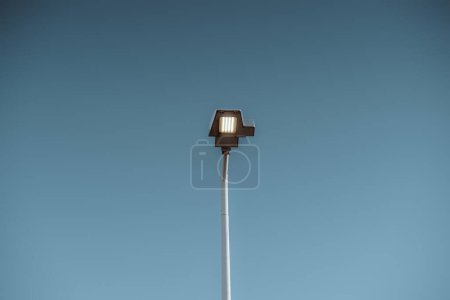 Téléchargez les photos : Une lumière d'inondation LED montée sur poteau à Lisbonne émet de la lumière pendant la lumière du jour, gaspillant de l'énergie sur un fond bleu clair - en image libre de droit