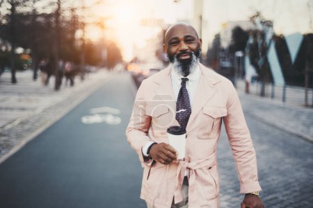 Téléchargez les photos : Un homme d'affaires noir chauve et barbu, vêtu d'une veste de couleur saumon, tient une tasse de café acheté tout en souriant d'oreille à l'oreille à Lisbonne, au milieu de la piste cyclable - en image libre de droit