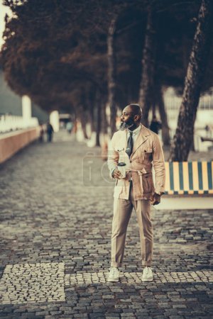 Téléchargez les photos : Un homme chauve et sophistiqué à barbe noire non rasée en pantalon beige et une veste regarde sur le côté tout en tenant un café à emporter sur un banc avec pavé de pavés à rayures jaunes et bleues à Lisbonne - en image libre de droit