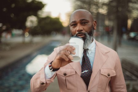 Photo for Lisbon; A bald man with a stylish beard poses for a close-up portrait; wears a pink jacket, white shirt, and polka dot tie. Holds a cup of coffee and is taking a sip; looks very charming and handsome - Royalty Free Image