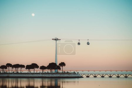 Foto de Lisboa; una hermosa escena de puesta de sol de bajo perfil con luna llena, puesta de sol de cielo rosa, el río Tejo y un teleférico. El efecto general es una escena mágica serena y de ensueño, con árboles ribereños - Imagen libre de derechos