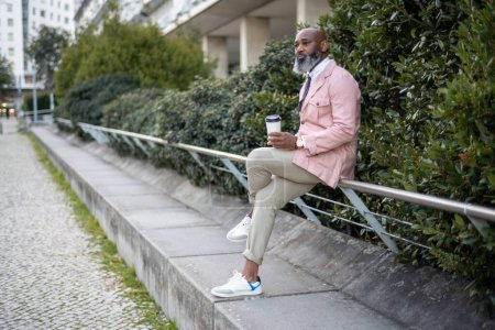 Photo for Lisbon; A black man with a bald head, wearing a pink jacket and a full-grown beard with white hair, holding a bought takeaway coffee cup while sitting on a railing resting against a shrubbery - Royalty Free Image
