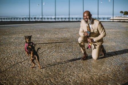 Téléchargez les photos : Lisbonne ; Un coup de feu d'un homme noir chauve avec une barbe promenant son chien sur le pavé pavé de Lisbonne avec un genou par terre, jauge le chien, tenant la laisse et son téléphone portable - en image libre de droit