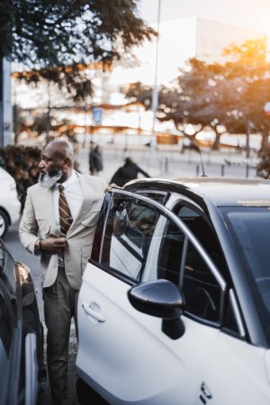 Photo for Lisbon; Vertical shot; Black male bald unshaven beard with white hairs fixing his jacket as he is about to enter his white car that is parked and adjacent to another vehicle - Royalty Free Image