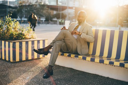 Photo for Lisbon; A well-dressed bald unshaven mature black man sitting on a wooden bench with yellow and blue vertical stripes; man checks the time on his wrist arm watch, his crossed legs showcase his ankles - Royalty Free Image