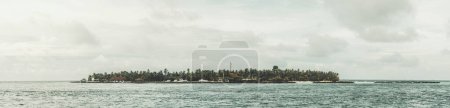 Photo for Maldives; Panorama of a Maldivian islet featuring lush green trees, surrounded by the vast calm blue sea, grey clouds in the sky contrast with the overall tranquil scene - Royalty Free Image