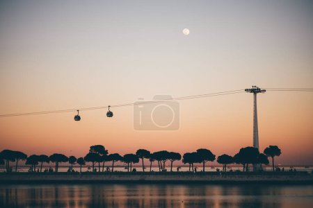 Téléchargez les photos : Une vue paisible et rêveuse de Lisbonne au crépuscule avec une pleine lune, un coucher de soleil rose, la rivière Tejo, deux télécabines et des silhouettes d'arbres au bord de la rivière - en image libre de droit