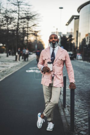 Photo for A stylish black bald man, with a long beard with hints of white hair, dressed in a pink jacket, holds a cup of coffee in the middle of a bicycle lane in Lisbon with a content expression on his face - Royalty Free Image