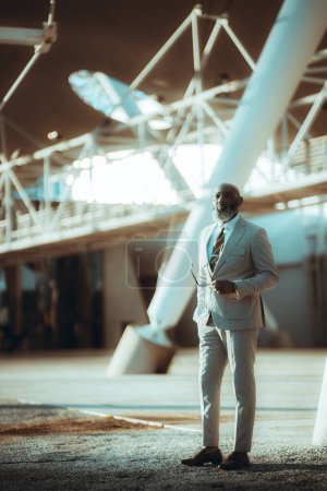Foto de Foto vertical de un hombre negro calvo de negocios con estilo, con una larga barba de pelo blanco, vestido con un traje, sostiene un par de gafas de sol con una expresión pensativa en un espacio arquitectónicamente hermoso - Imagen libre de derechos
