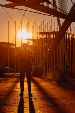 Photo for Vertical; In Lisbon, a black man stands before a white tensile structure, bathed in golden hour light. Dramatic backlighting creates captivating long shadows, setting a low-key and atmospheric scene - Royalty Free Image