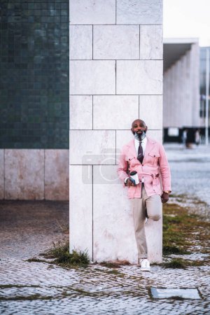 Foto de Un hombre calvo negro elegante, con una larga barba gris, vestido con una chaqueta de abrigo rosa, sostiene una taza de café mientras se apoya sobre un pilar de mármol blanco con una expresión atractiva en su cara - Imagen libre de derechos