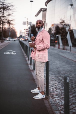 Foto de Un hombre calvo negro con estilo, con una larga barba de pelo gris, vestido casualmente de rosa, sostiene una taza de café mientras está a punto de cruzar el carril bici por la tarde, comprueba si el camino está despejado - Imagen libre de derechos