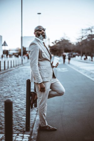Photo for A vertical capture of a stylish black bald man, with an unshaven white-haired beard, wearing a beige suit, holds a cup of coffee while waiting against a post delimiting the bicycle path in Lisbon - Royalty Free Image
