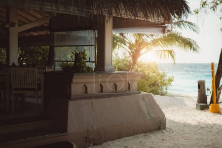 Photo for Sunny day at a sandy beach cafe made of sandstone with a thatched roof. Palm trees surrounding it can be seen in the back, in front of the sea, providing an exotic tranquil vibes - Royalty Free Image