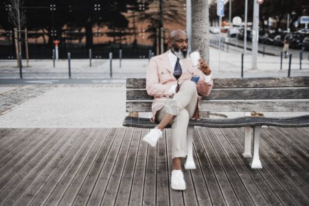 Foto de Tiro vertical: Un hombre negro calvo bien vestido sentado en un banco de madera con una taza de café cerca. Su chaqueta de melocotón, pantalones de crema y teléfono en la mano reflejan su comportamiento seguro y elegante - Imagen libre de derechos