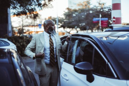 Photo for In the streets of Lisbon, a confident black man with a bald head and a long beard adjust his cream suit jacket with precision. Standing beside his white car, parked adjacent to another vehicle - Royalty Free Image
