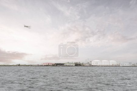 Foto de Maldivas: aguas grises del mar y cielo nublado en primer plano, el aeropuerto de Male exhibe infraestructura vital con tanques de combustible y tanques de almacenamiento en el fondo - Imagen libre de derechos