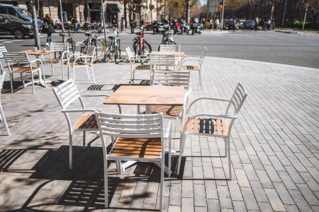Foto de Barcelona; Amplio espacio en la acera adornado con mesas y sillas vacías, atractivas, blancas y marrones, perfectamente espaciadas para que los visitantes disfruten de momentos mientras las bicicletas descansan cerca, un lugar cautivador para relajarse - Imagen libre de derechos
