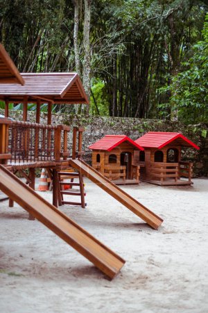 Foto de Una toma vertical de un parque infantil en un entorno tropical con un enfoque selectivo en las dos pequeñas casas de madera con techos de plástico rojo para niños en la arena; dos toboganes para niños en un primer plano desenfocado - Imagen libre de derechos