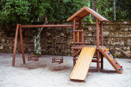 Foto de Un parque infantil en un parque tropical con dos columpios de cadena infantil y un tobogán de madera para niños en la arena con un techo triangular de la construcción y repisas multicolores de las escaleras - Imagen libre de derechos