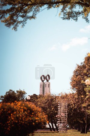 Foto de Telefoto medio vertical de un hermoso parque en colores cálidos de verano y otoño con enfoque selectivo en la torre en el fondo con dos anillos de un Monumento 25 de Abril en Lisboa, Portugal - Imagen libre de derechos