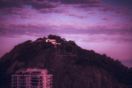 Téléchargez les photos : Plan à long foyer d'une montagne du soir au crépuscule avec un fort sur le dessus et le toit d'un bâtiment résidentiel au premier plan dans des couleurs modernes toxiques rose et violet avec un ciel partiellement nuageux au-dessus - en image libre de droit