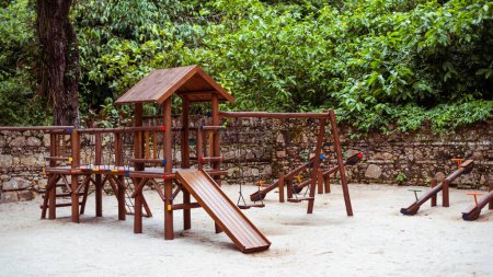 Foto de Vista de un parque infantil en un parque tropical con un grupo de balancines en el fondo junto a la pared de piedra, tobogán de madera para niños y columpio frente a los tambaleantes en la arena - Imagen libre de derechos
