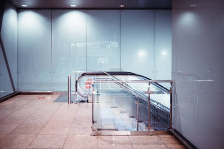 Téléchargez les photos : Vue d'un escalier roulant et d'un escalier moderne en verre et chrome dans un terminal aéroportuaire contemporain ou un centre commercial ou une station de métro ; sol carrelé et murs en verre reflétant l'intérieur du passage - en image libre de droit