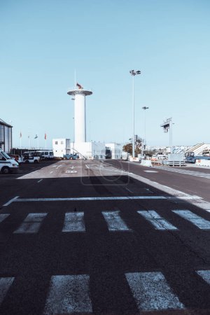 Photo for Vertical shot of an outdoor airport area with a VOR (VHF Omnidirectional Range) antenna tower, an asphalt road in a shadow with a pedestrian crossing, and the runway maintenance infrastructure - Royalty Free Image