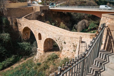 Photo for Wide angle outdoor view of two bridges over a dry channel depression lit by the sun: a stone arch pedestrian bridgeand a concrete one for transport; stairs in the foreground; Caldes de Montbui, Spain - Royalty Free Image