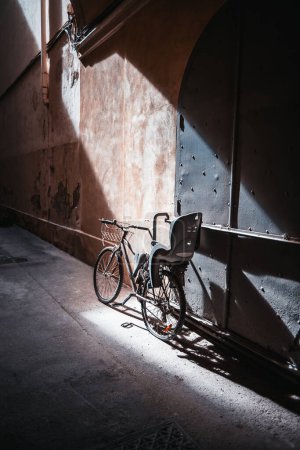 Photo for Vertical contrast shot of an old bicycle with an extra child seat and basket lit by the sun and parked next to a flaked wall and riveted metal gates in a shadow of a narrow street in Barcelona, Spain - Royalty Free Image