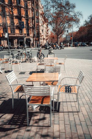 A vertical shot of a sunlit outdoor cafe setting in a bustling Barcelona city with empty tables and chairs, inviting a moment of urban relaxation