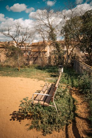 A vertical shot with a shallow depth of field and selective focus on a rustic urban bench overlooks a tranquil park path next to a playground with a clay floor