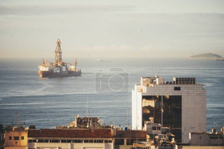 An offshore oil rig stands majestically in the open sea at dawn, viewed from Leme, Rio. The rig, bathed in the light, contrasts with the urban skyline, highlighting the blend of industry and nature