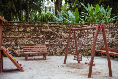 Ruhiger Kinderspielplatz inmitten eines tropischen Parks mit Holzbank, Schaukel und farbenfroher Kletterstruktur. Üppiges Laub und eine historische Steinmauer schaffen eine heitere Kulisse