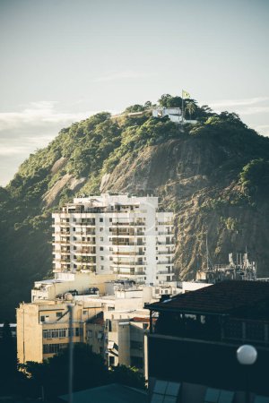 Leme's distinct urban landscape, lush green hill topped with a historic fort, juxtaposed against modern residential buildings in the foreground. A blend of Rio's natural and urban elements