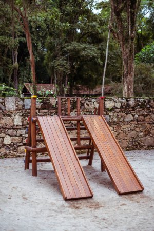 Captured in a lush tropical park, this children's playground features wooden slides and swings, set against a backdrop of thick greenery and a stone wall, offering a natural and safe play area