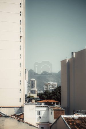 Urban cityscape of Rio de Janeiro featuring modern high-rise buildings framed between tall residential structures. Distant mountains provide a scenic backdrop under a clear sky, vertical shot