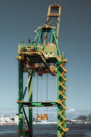 Close-up view of a large industrial crane at a port, with yellow and green structure against a clear blue sky. The background features a coastal city skyline and mountains. Good for themes of industry