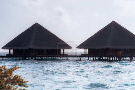 Deux bungalows traditionnels au-dessus de l'eau avec des toits de chaume situés sur pilotis au-dessus des eaux turquoise des Maldives. La scène capture la tranquillité et la beauté de la vie insulaire des escapades tropicales