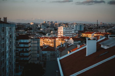 View of Almada cityscape at sunset featuring a mix of residential buildings with red-tiled roofs and modern high-rises. The warm sunlight highlights the architecture against a backdrop of urban sprawl