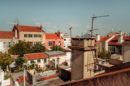 Paisaje urbano de Lisboa con edificios residenciales tradicionales con techos de tejas rojas, antenas visibles y exuberantes jardines. Un cielo azul claro y la luz del sol resaltan el barrio urbano.