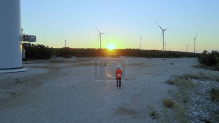 Photo for Male Technician In Helmet Walking In The Field Towards The Wind Turbines At Sunset - Royalty Free Image