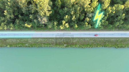 Red electric car driving on straight road with pine tree forest and water lake. Graphic logo of battery charging, aerial top down