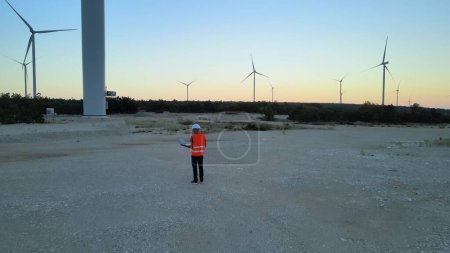 Antenne eines Ingenieurs mit Laptop, der die Effizienz des Windkraftparks bei Sonnenuntergang inspiziert und kontrolliert, klares Konzept für erneuerbare Energien mit null Emissionen