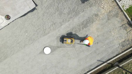 Luftaufnahme über einem Mann, der Fundament mit Vibrationsplatte stampft - von oben nach unten, Drohnenschuss
